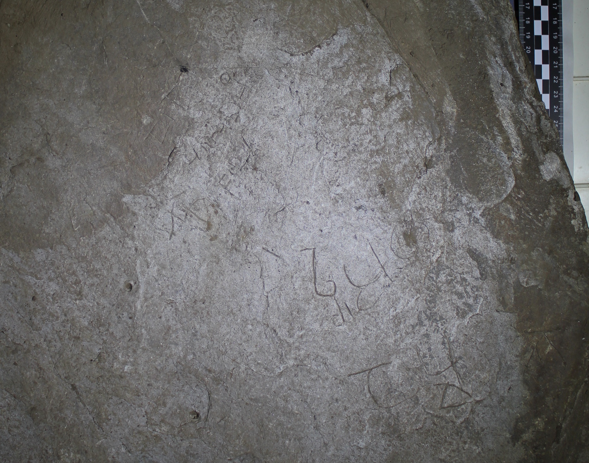 An overhead image of a large lump of slate with a reasonably flat surface. Just about visible are some faint letters incised into the slate, made slightly clearer by the shadows created from the directional light source.