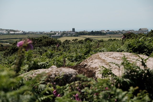 View over the two remaining capstones of Tregeseal Entrance Grave in the foreground. Green ferns and pink foxgloves cover the lower half of the image. In the background the houses, chapels and church tower in St Just are visible with its parapets, and the hazy sea is beyond.