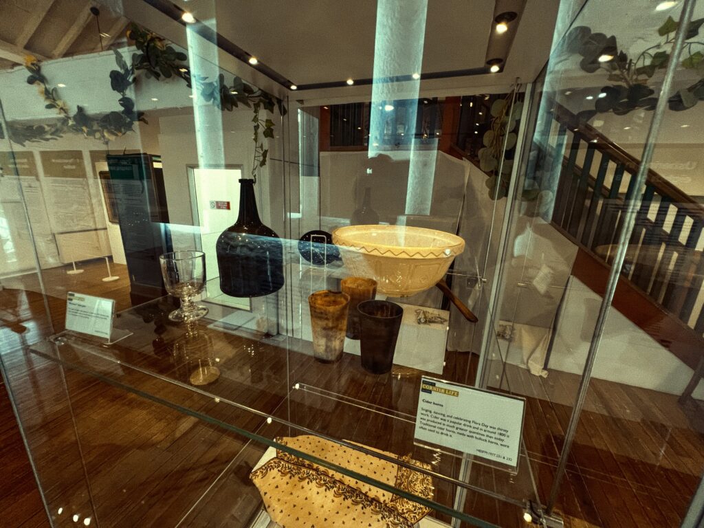 The second image shows another angle of the exhibition, focusing on a different glass display case. This case contains various objects: a large blue vase, a clear glass with intricate designs, a wooden bowl, and a yellow patterned ceramic bowl. In the foreground, there’s a yellow textile with brown patterns. Descriptive labels are present but not legible in the photo. A stair banister on the right and an informational panel in the back hint at the layout of the museum. The ambiance remains warm, with wooden floors and soft lighting.