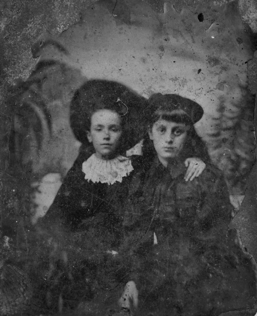 A very old photograph from the 1860s. Two young people, possibly a boy and a girl sitting side by side looking at the camera. They are both wearing hats and mid 19th century clothes.