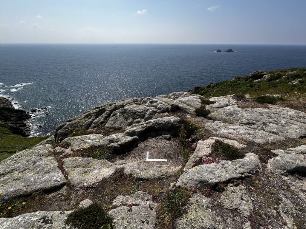 The setting of the grey granite of Carn Leskys on the clifftop, with the sea and blue sky, and the two islets of the Brisons in the distance on the right-hand side.