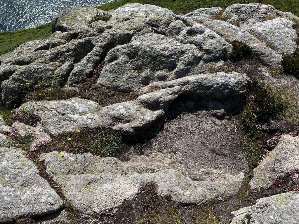 A view of the granite stone containing prehistoric motifs. Much of the grey granite is covered in spiky pale green lichen. Pockets of heather and grass can be seen. The sea below the cliffs is just visible indicating the height of this rock above the cliffs.