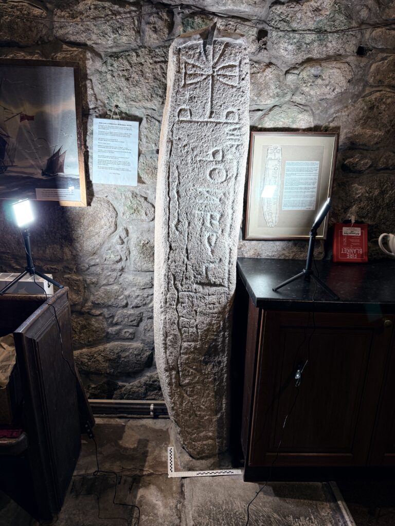 Tall early medieval memorial stone standing inside Madron church against a rough granite wall, carved with a long vertical inscription and topped by a simple cross with flared arms.