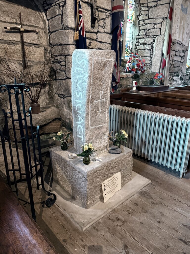 Early medieval inscribed stone standing inside a stone church, with an ancient vertical inscription carved on one face and a tau-rho symbol incised on the adjacent face, mounted on a square base beside wooden pews, flags, flowers and a radiator.