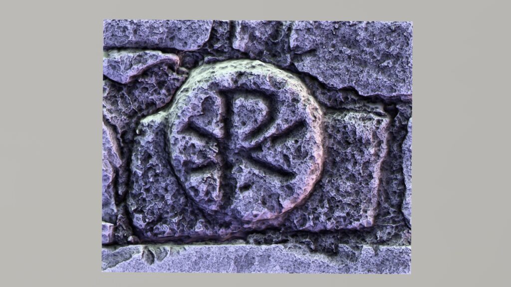 Close-up of a weathered medieval chi-rho symbol carved in stone, with the overlapping Greek letters Chi and Rho enclosed in a circular form and set into a rough stone wall.